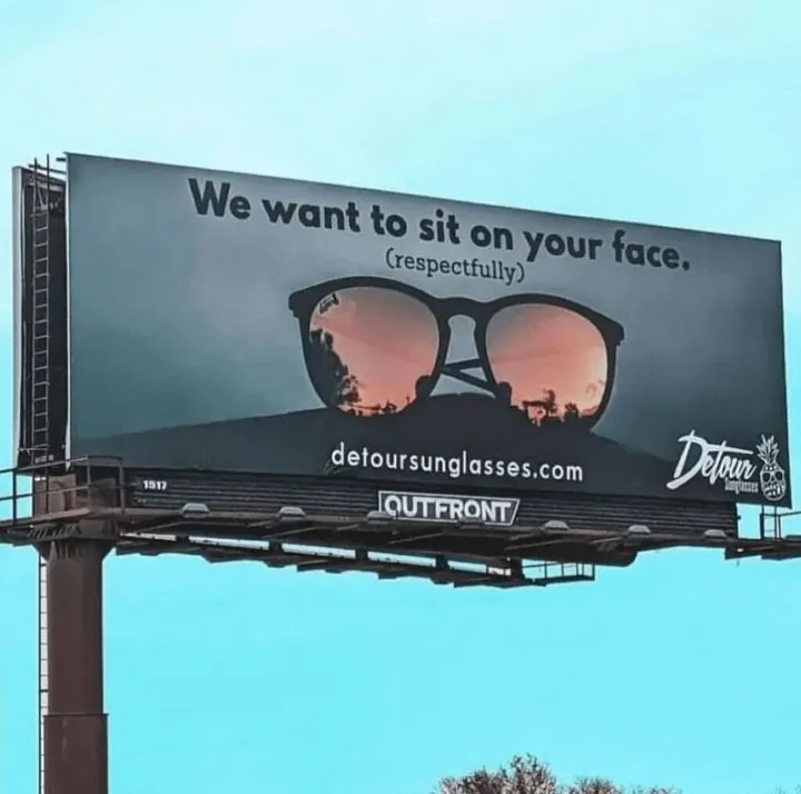 Detour Sunglasses billboard against a blue sky reading 'We want to sit on your face. (respectfully)' above sunglasses reflecting a sunset.